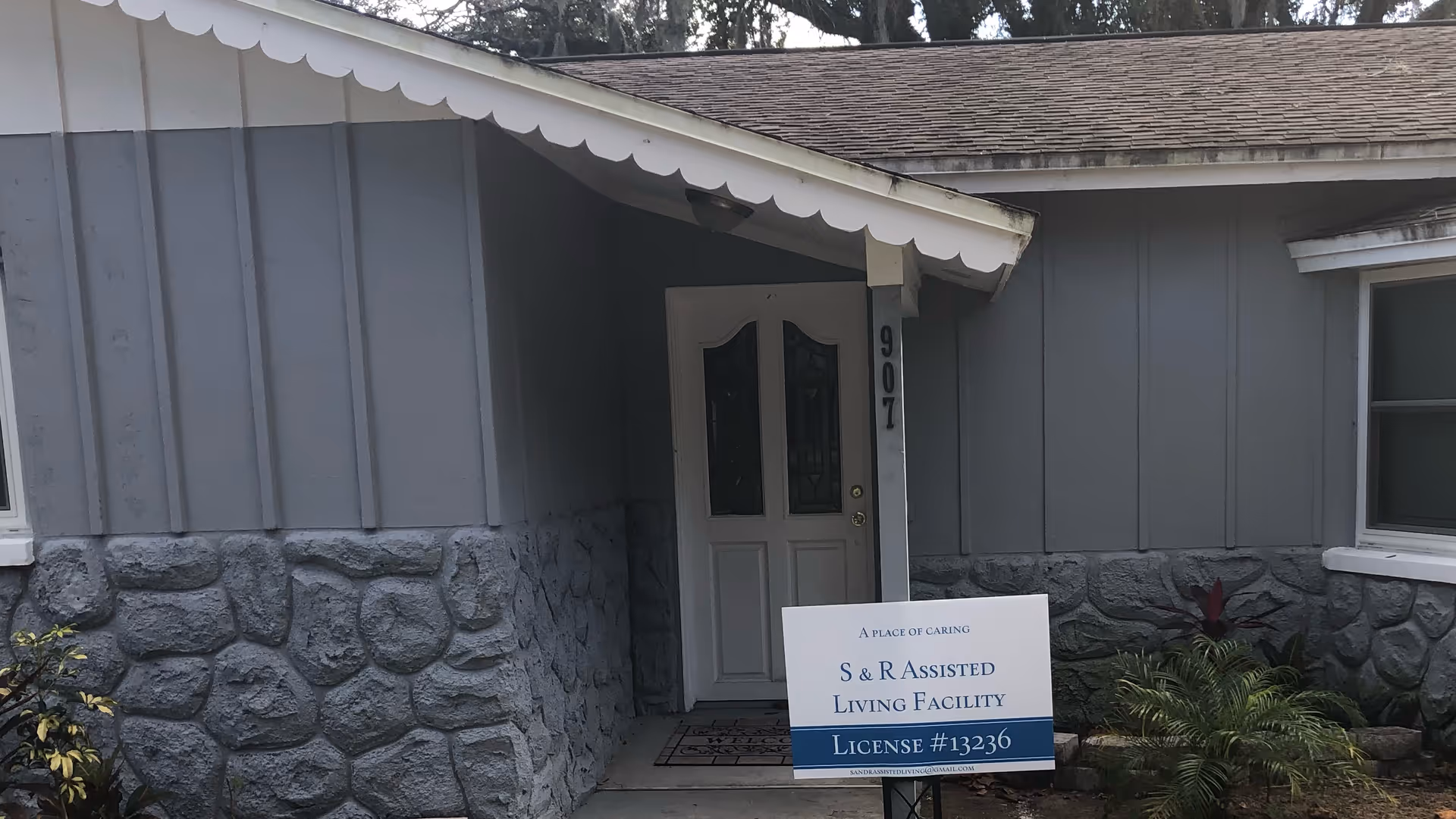 Front entrance of a gray single-story building with a white door and a lawn sign reading 'S & R Assisted Living Facility, License #13236'.