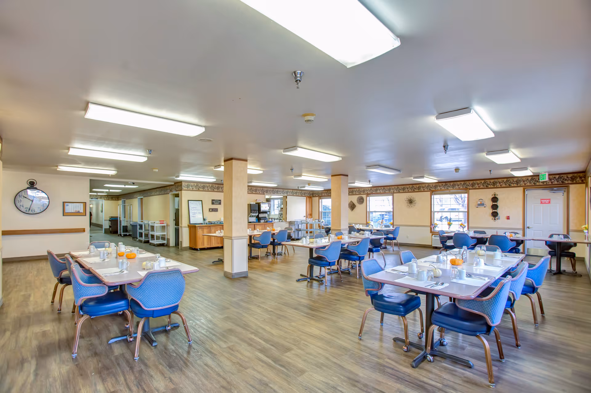 A spacious dining room in a senior living facility with multiple tables set for meals. Each table has blue chairs around it and is arranged with white cups, plates, and utensils. The room has large windows letting in natural light, wood flooring, and fluorescent ceiling lights. There is a coffee station and a clock on the wall.