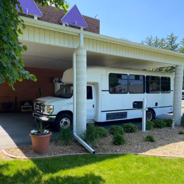 A white shuttle bus parked under a covered driveway area next to a building with a brick wall and purple roof accents. There are plants and a flower pot near the driveway and green grass in the foreground.