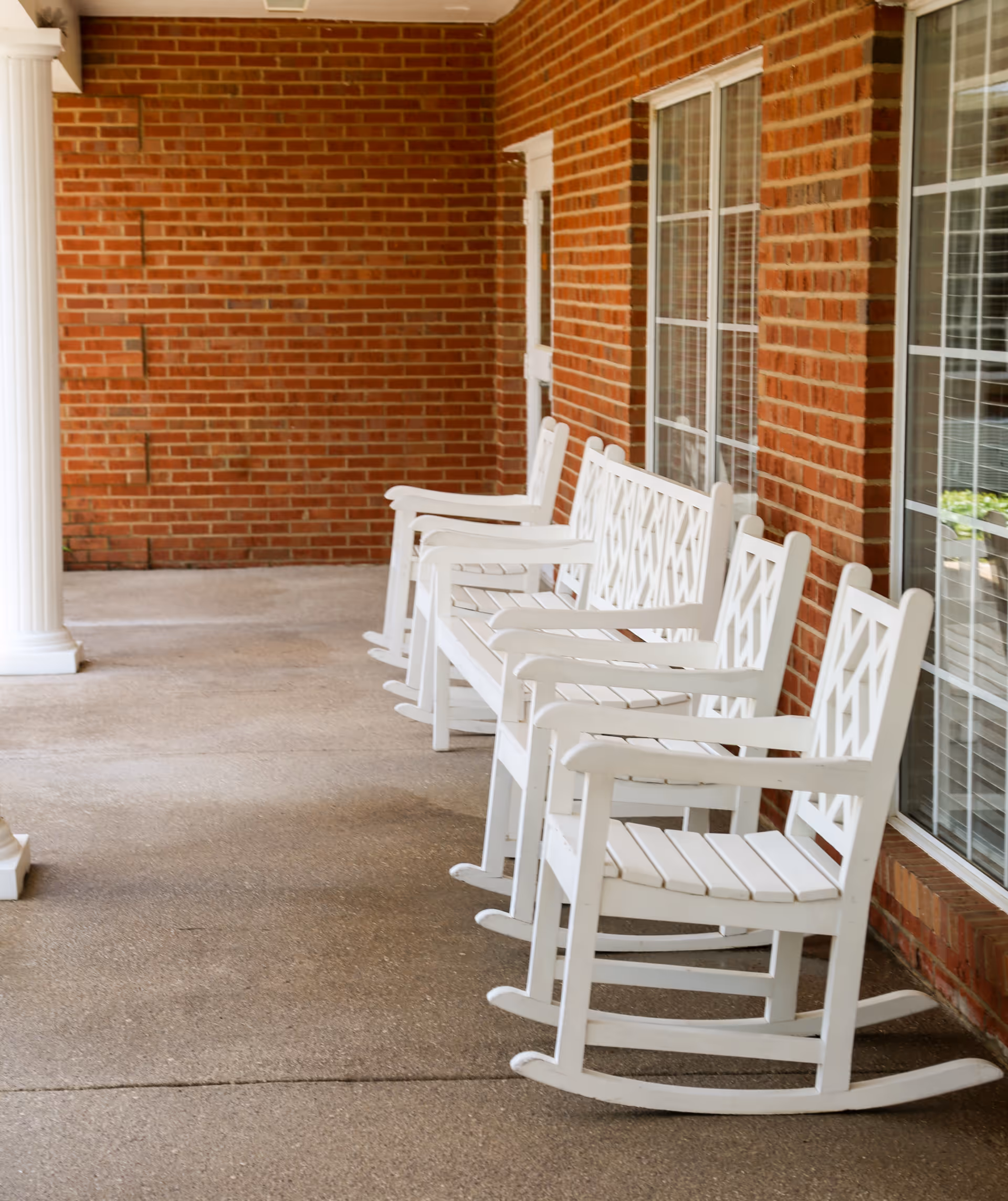 A row of white wooden rocking chairs on a covered brick-front porch next to windows.