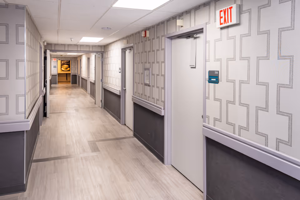 A clean, well-lit hallway in a nursing and rehabilitation center with patterned wallpaper, light wood flooring, and several closed doors along the walls. An exit sign is visible above one door, and a sign with the number 206 is mounted next to it. At the end of the hallway, there is a sign with a coffee cup image and the word 'ROAST'.