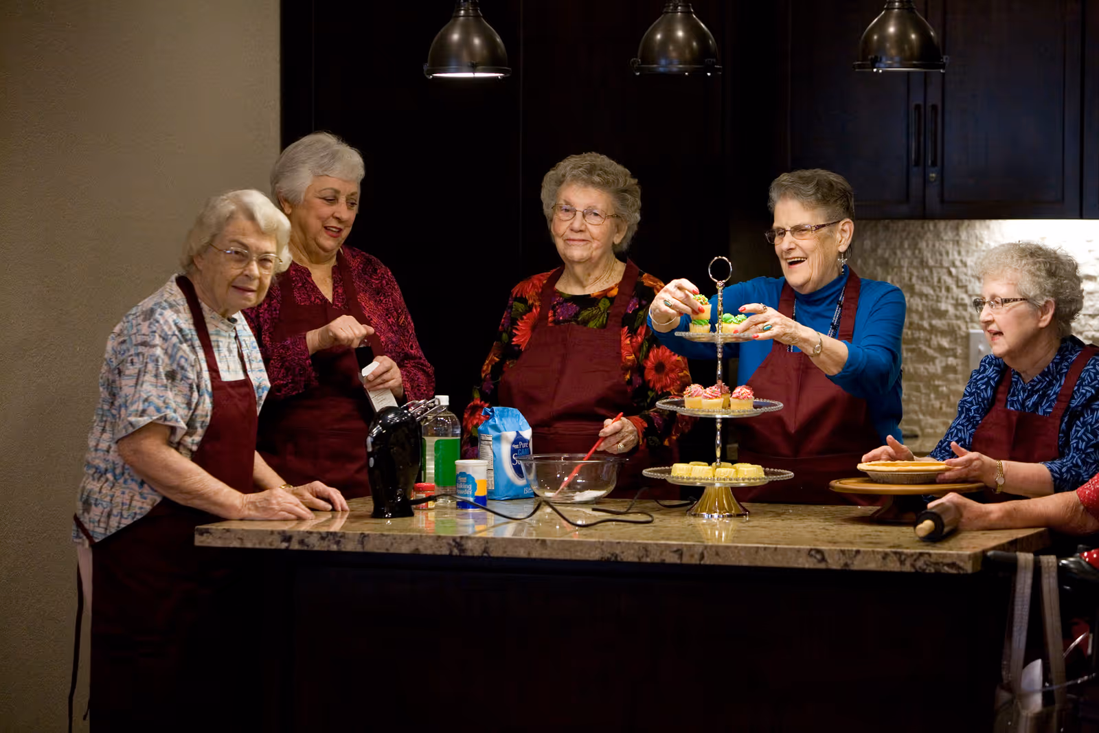 Five elderly women wearing aprons are gathered around a kitchen island, preparing and arranging baked goods such as cupcakes and pies. The setting appears to be a warm, well-lit kitchen with dark cabinets and hanging pendant lights.