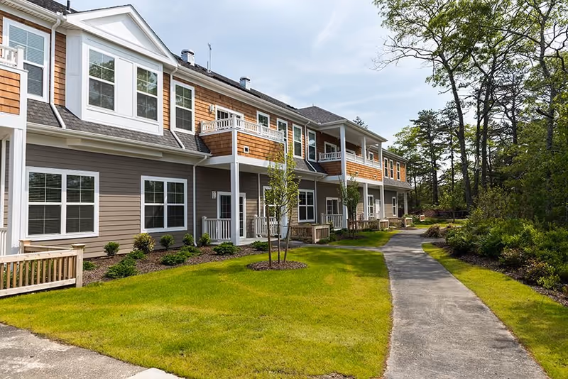 Exterior view of a two-story residential building with a combination of brown and gray siding, white trim, and multiple windows. The building has small balconies and porches, surrounded by a well-maintained lawn, young trees, and a paved walkway leading alongside the building. Trees and greenery are visible in the background under a partly cloudy sky.