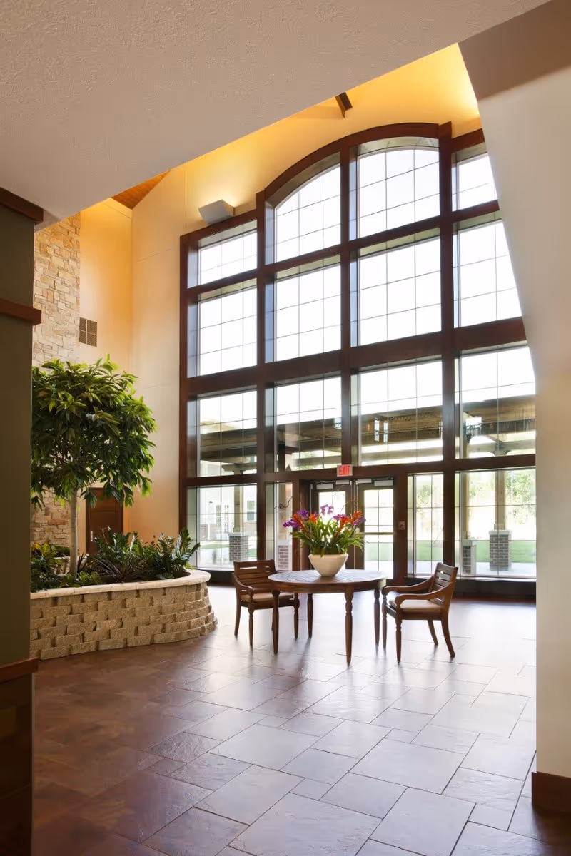 A bright interior space with large floor-to-ceiling windows featuring a grid pattern and an arched top. In front of the windows is a round wooden table with a white vase filled with colorful flowers. Two wooden chairs are placed on either side of the table. To the left, there is a raised planter with green plants and a small tree. The floor is tiled with large brown tiles, and the walls are painted in warm tones with a stone accent wall visible on the left.
