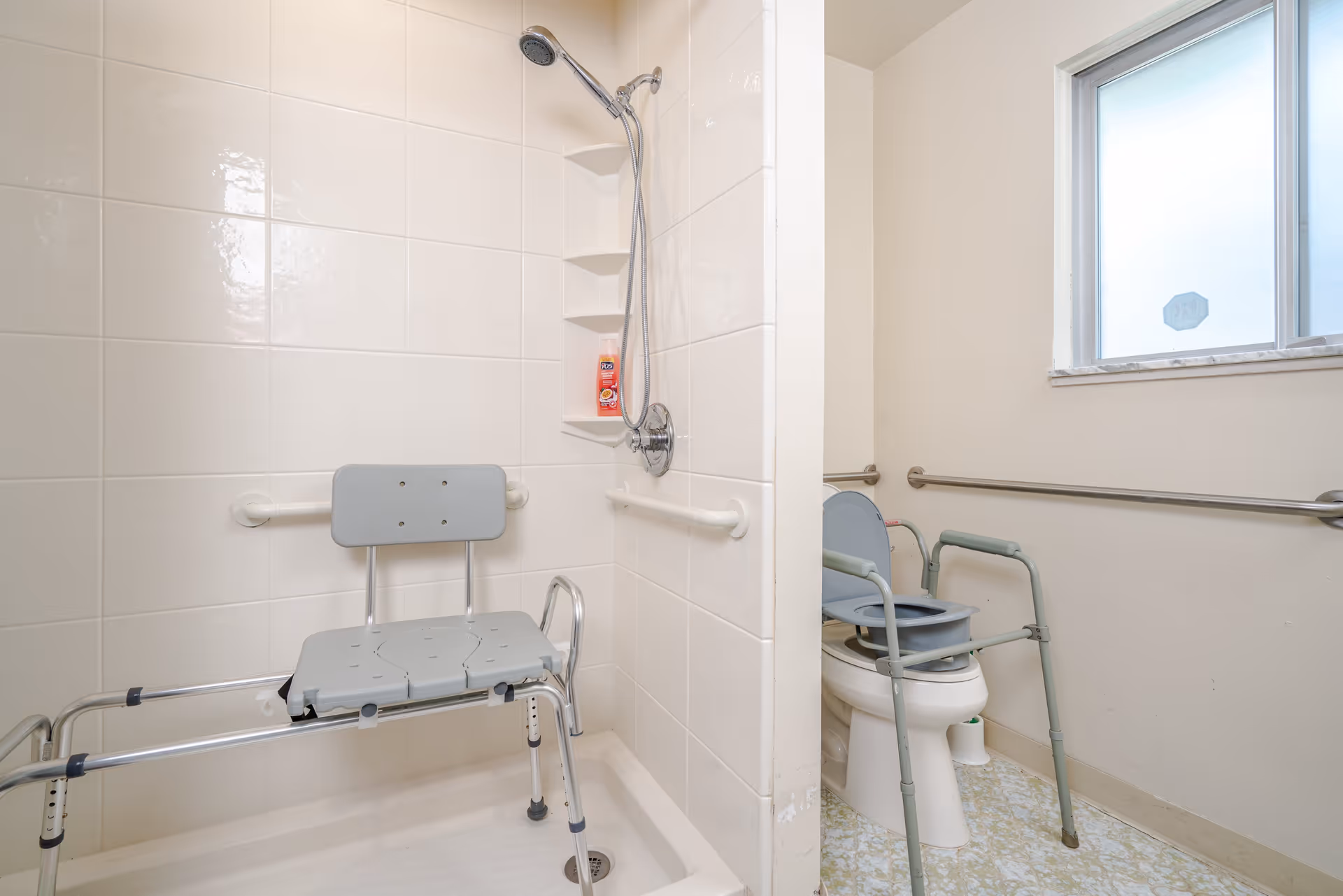 A bathroom designed for accessibility featuring a shower area with a shower chair and grab bars, and a toilet equipped with a raised seat and a walker nearby. The room has tiled walls in the shower and a frosted window providing natural light.