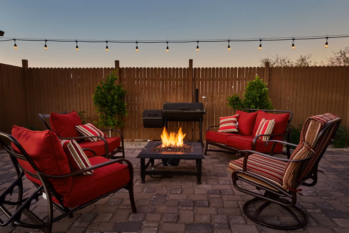 Outdoor patio area with a fire pit in the center surrounded by four cushioned chairs and a loveseat with red and striped pillows. String lights hang above, and there is a wooden fence and some greenery in the background.
