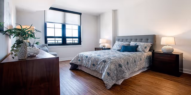 A bright bedroom with a large window, a bed with patterned bedding and pillows, two bedside tables each with a lamp, a wooden dresser with plants and decorative items, and wooden flooring.