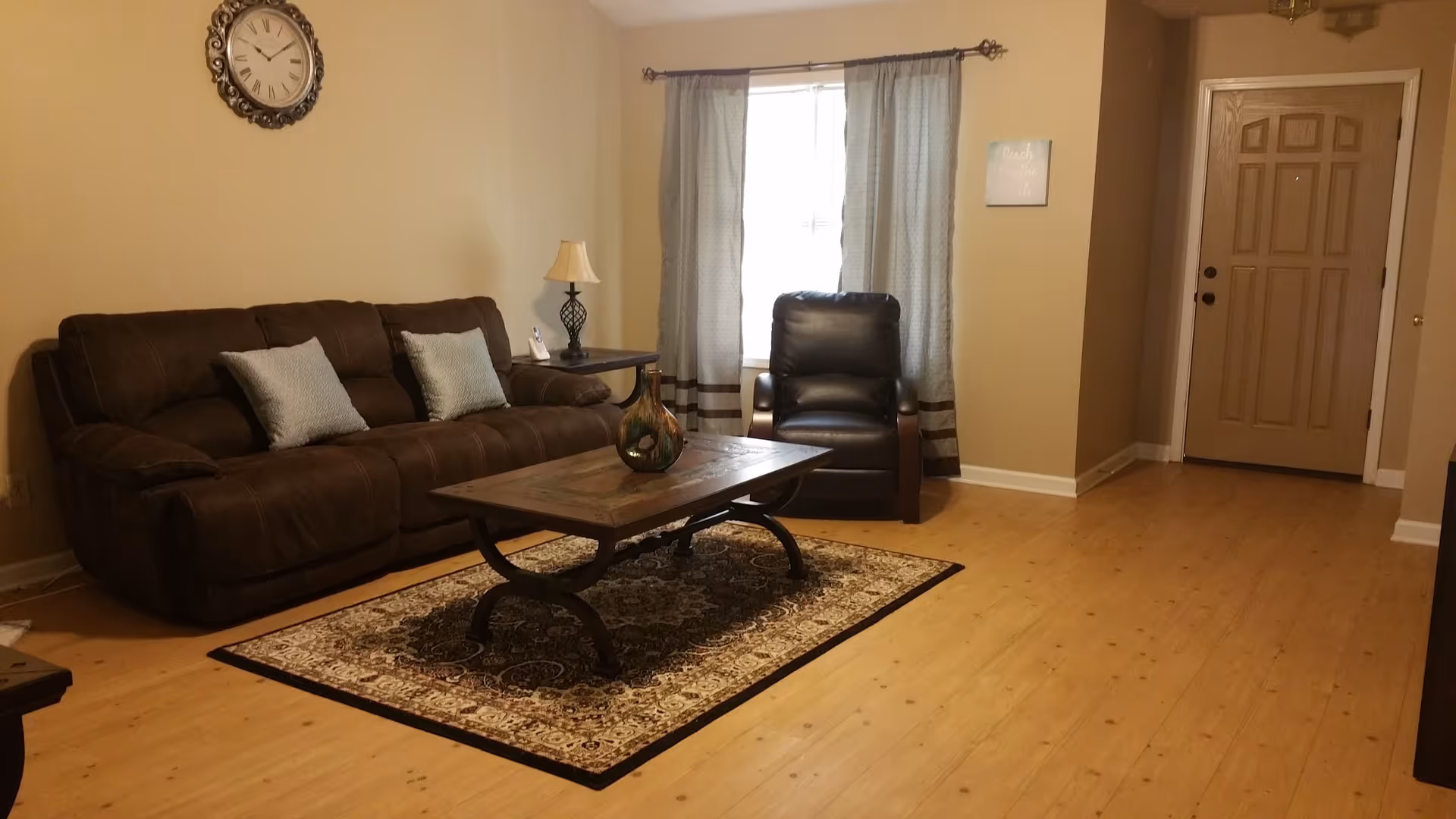 A cozy living room with a brown sofa, two light blue cushions, a dark brown leather armchair, a wooden coffee table with a decorative vase, a patterned area rug, a side table with a lamp, a wall clock, and a window with light-colored curtains. The room has light wood flooring and beige walls, with a closed wooden door in the background.