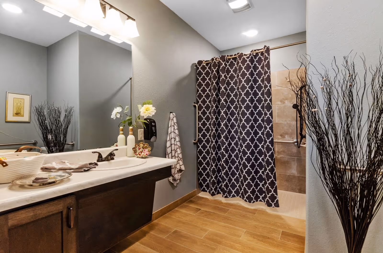 A bathroom with a large mirror above a sink with a dark wood cabinet. The sink countertop has decorative items including two white vases with flowers and a small dish. There is a towel hanging on the wall next to a soap dispenser. The shower area has a dark patterned curtain and beige tiled walls with grab bars. The floor is wood-like tile, and there are decorative tall twigs in a vase on the right side.
