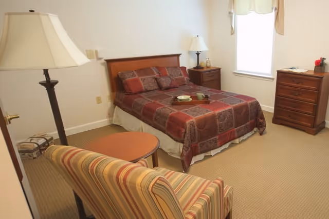 Furnished bedroom with a double bed, matching bedside tables and lamps, a striped armchair, small table, and a dresser under a window.