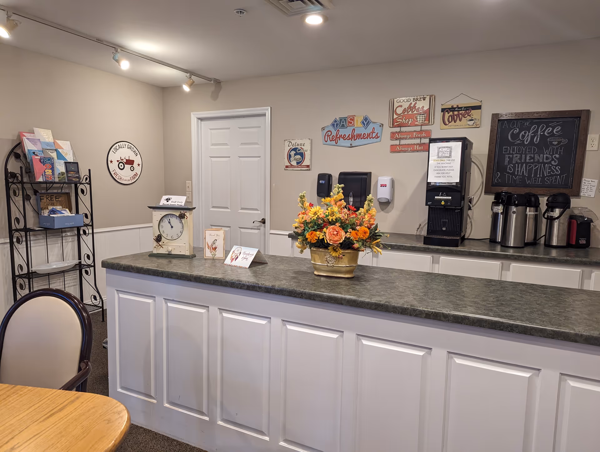 Interior view of a refreshment area in a senior living facility with a countertop featuring a flower arrangement, a clock, and small signs. Behind the counter are coffee dispensers, a coffee machine, and wall decorations including signs about coffee and refreshments. A metal rack with brochures and a chair next to a wooden table are also visible.
