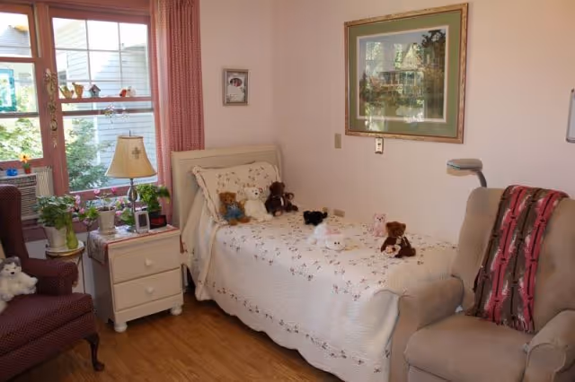 Cozy bedroom with a single bed covered by a white quilt and stuffed animals, a nightstand with a lamp and plants, and armchairs by a window.
