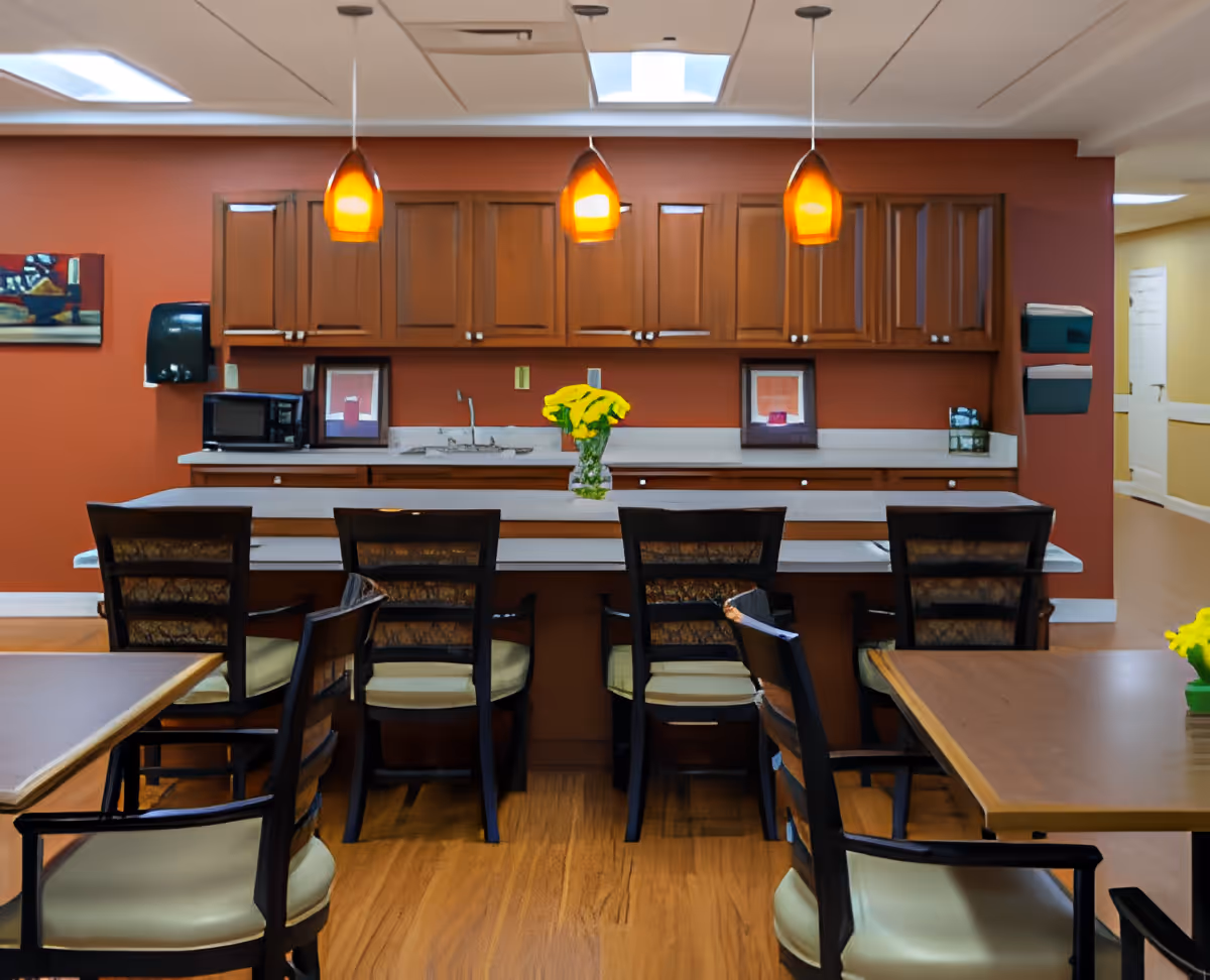 Interior view of a dining area with a kitchen counter and bar stools. The counter has a vase with yellow flowers, and above it hang three pendant lights with orange shades. Wooden cabinets are mounted on the wall behind the counter, and there are framed pictures and a microwave on the countertop. Several dining tables and chairs are visible in the foreground.