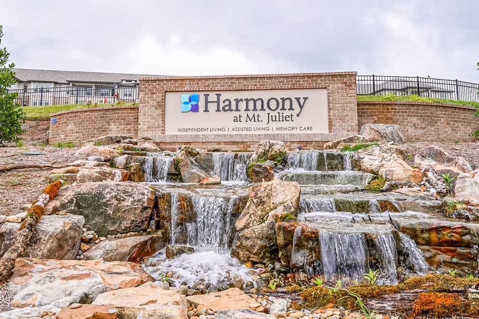 Outdoor view of a stone waterfall feature in front of a brick sign for Harmony at Mt. Juliet, a senior living facility offering independent living, assisted living, and memory care. The sign is surrounded by rocks and greenery with a cloudy sky in the background.