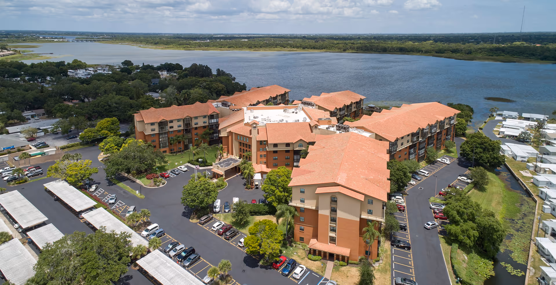 Aerial view of Lake Seminole Square senior living facility with multiple connected buildings featuring terracotta roofs, surrounded by parking lots, trees, and a large body of water in the background under a partly cloudy sky.
