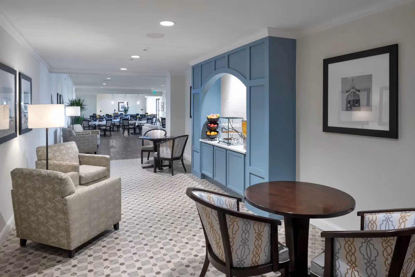A bright and spacious common area in a senior living facility featuring comfortable armchairs and small round tables with patterned chairs. The floor has a decorative tile pattern, and there is a blue built-in nook with a countertop holding fruit and beverages. In the background, there is a dining area with multiple tables and chairs, illuminated by ceiling lights.