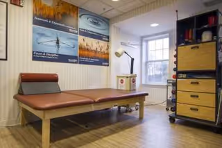 A therapy or treatment room with a padded examination table, a lamp, a window letting in natural light, and a shelving unit with drawers and various items. The walls have motivational posters related to wellness and therapy.