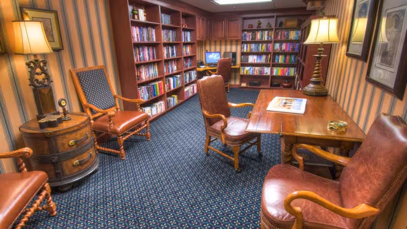 A cozy library room with wooden bookshelves filled with books along the walls, a wooden table with leather chairs around it, two table lamps providing warm lighting, and a computer desk with a chair in the back corner. The room has patterned carpet and striped wallpaper.