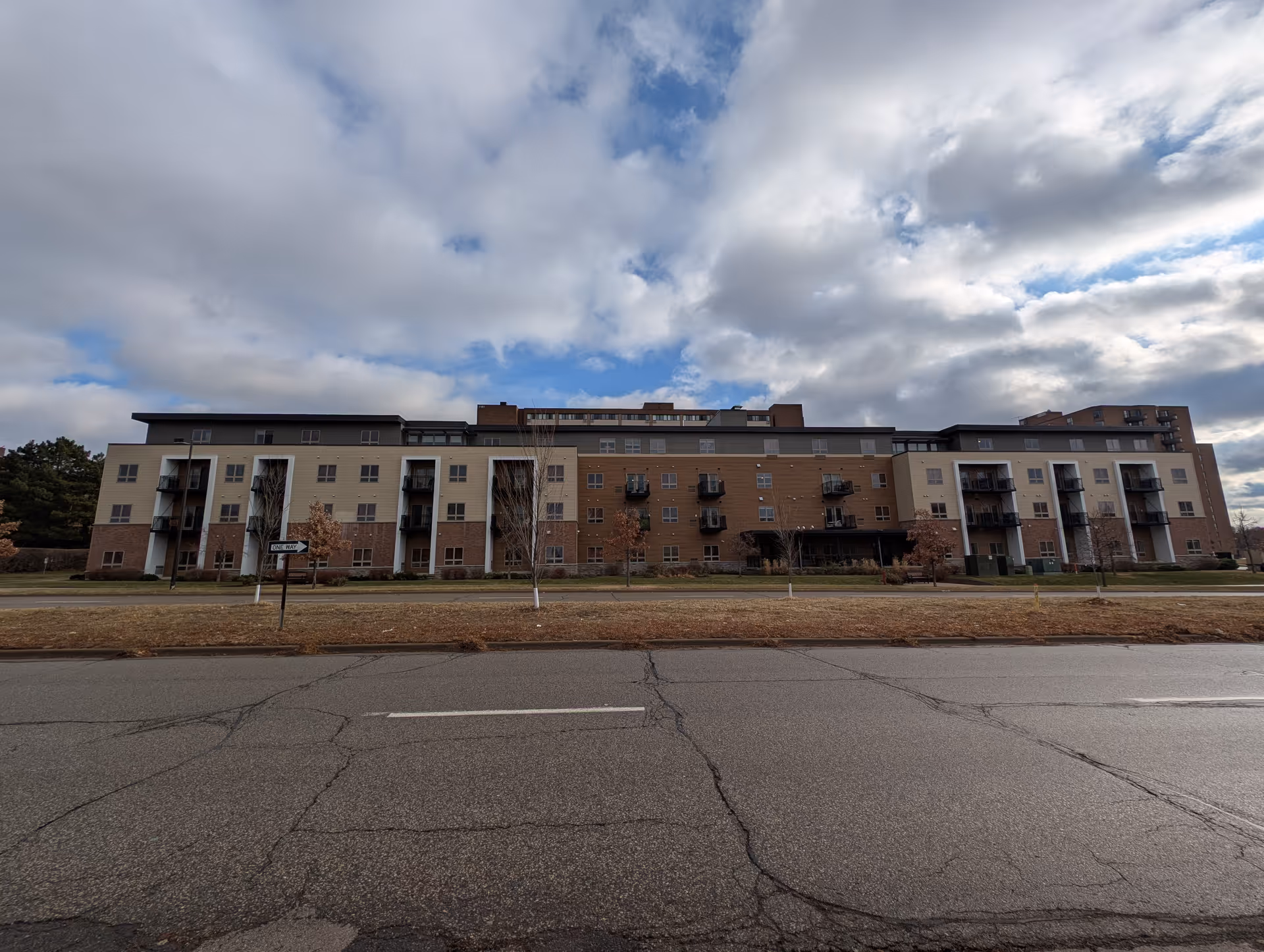 Exterior view of a multi-story senior living facility building with balconies, set against a partly cloudy sky. The building is surrounded by a grassy area with some small trees and a road in the foreground.