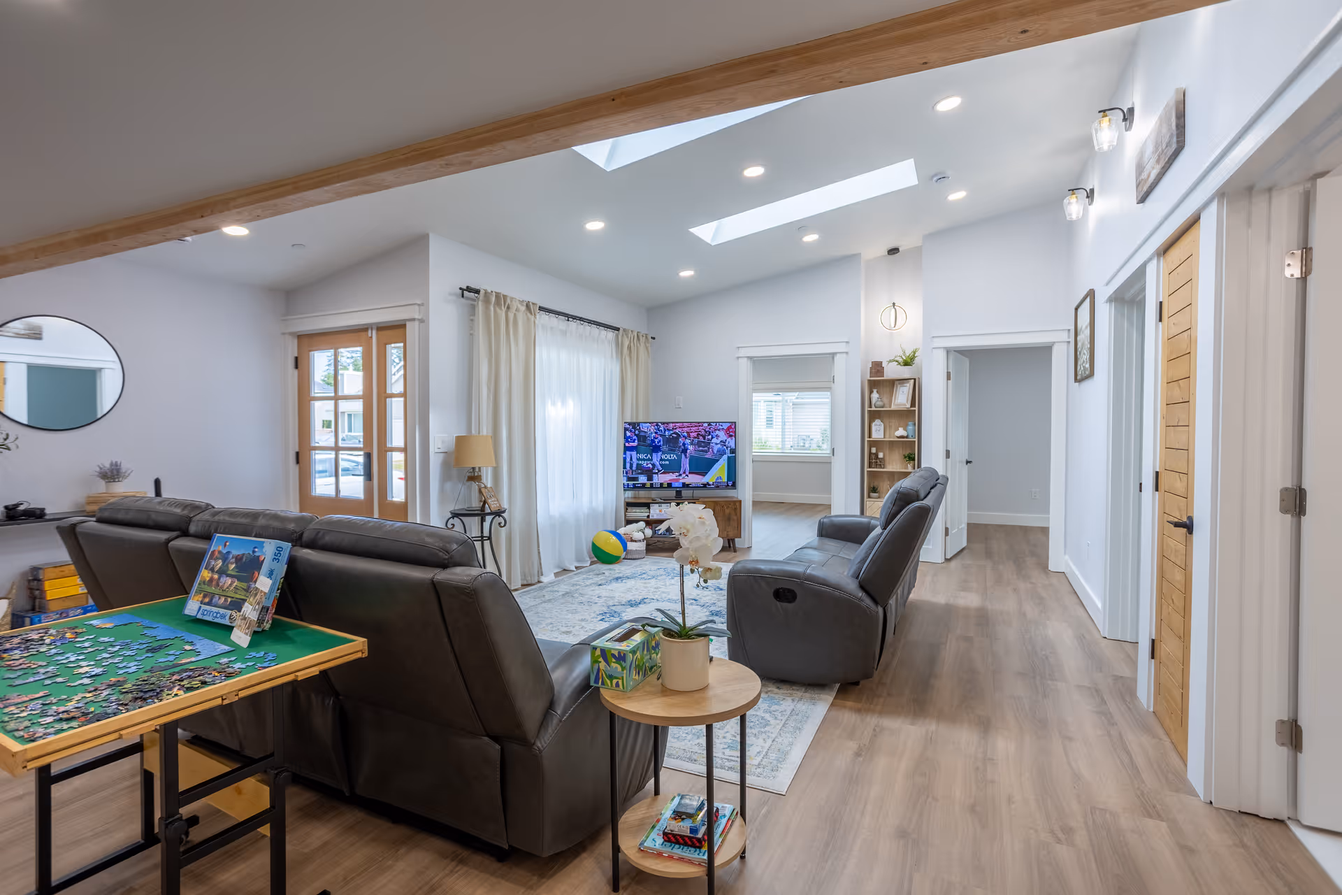 Bright modern living room with leather recliners, a TV, skylights, and a puzzle table in the foreground.