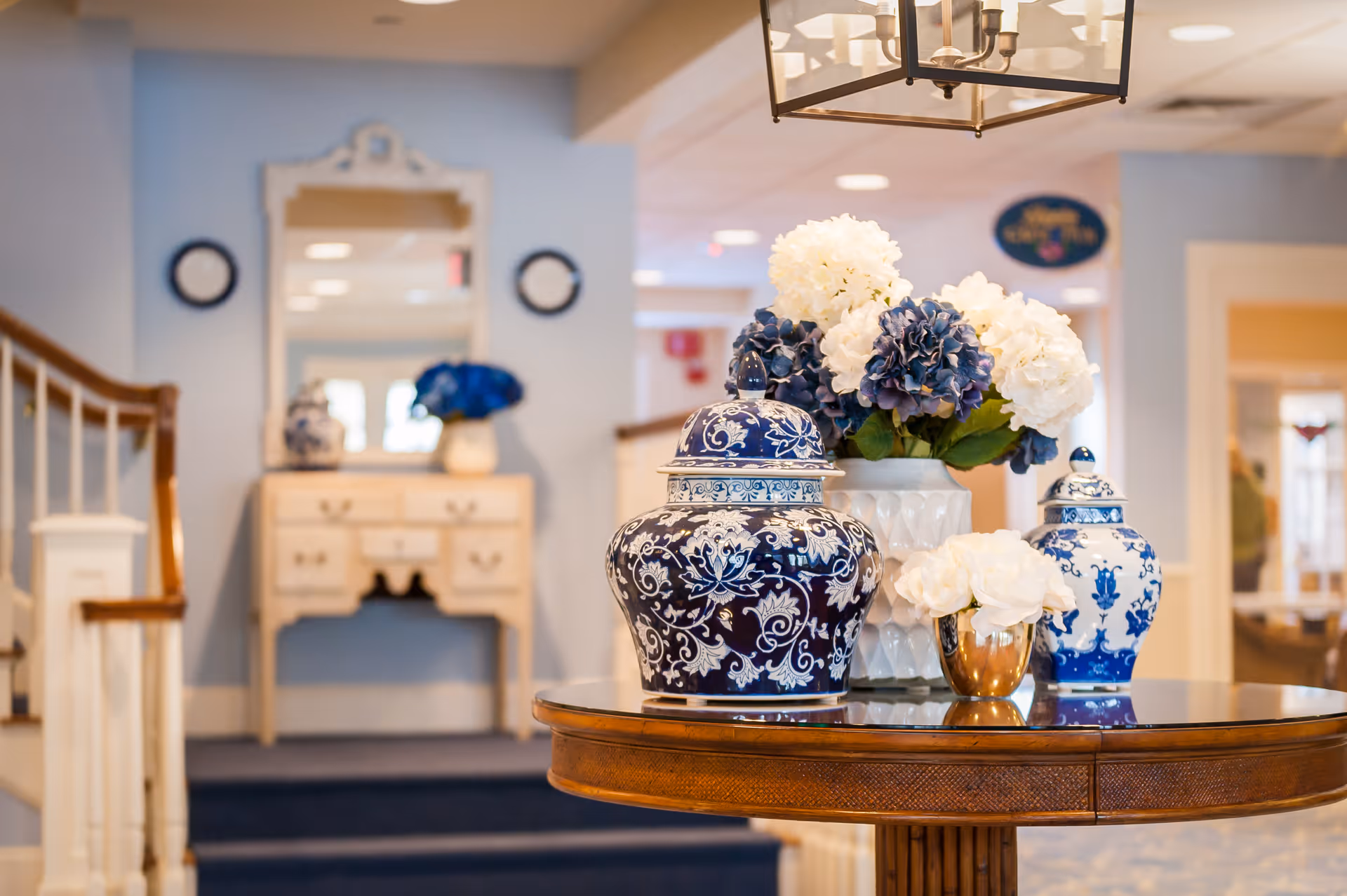 Interior foyer with a round wooden table displaying blue-and-white ceramic vases and floral arrangements, with a staircase and console table in the background.