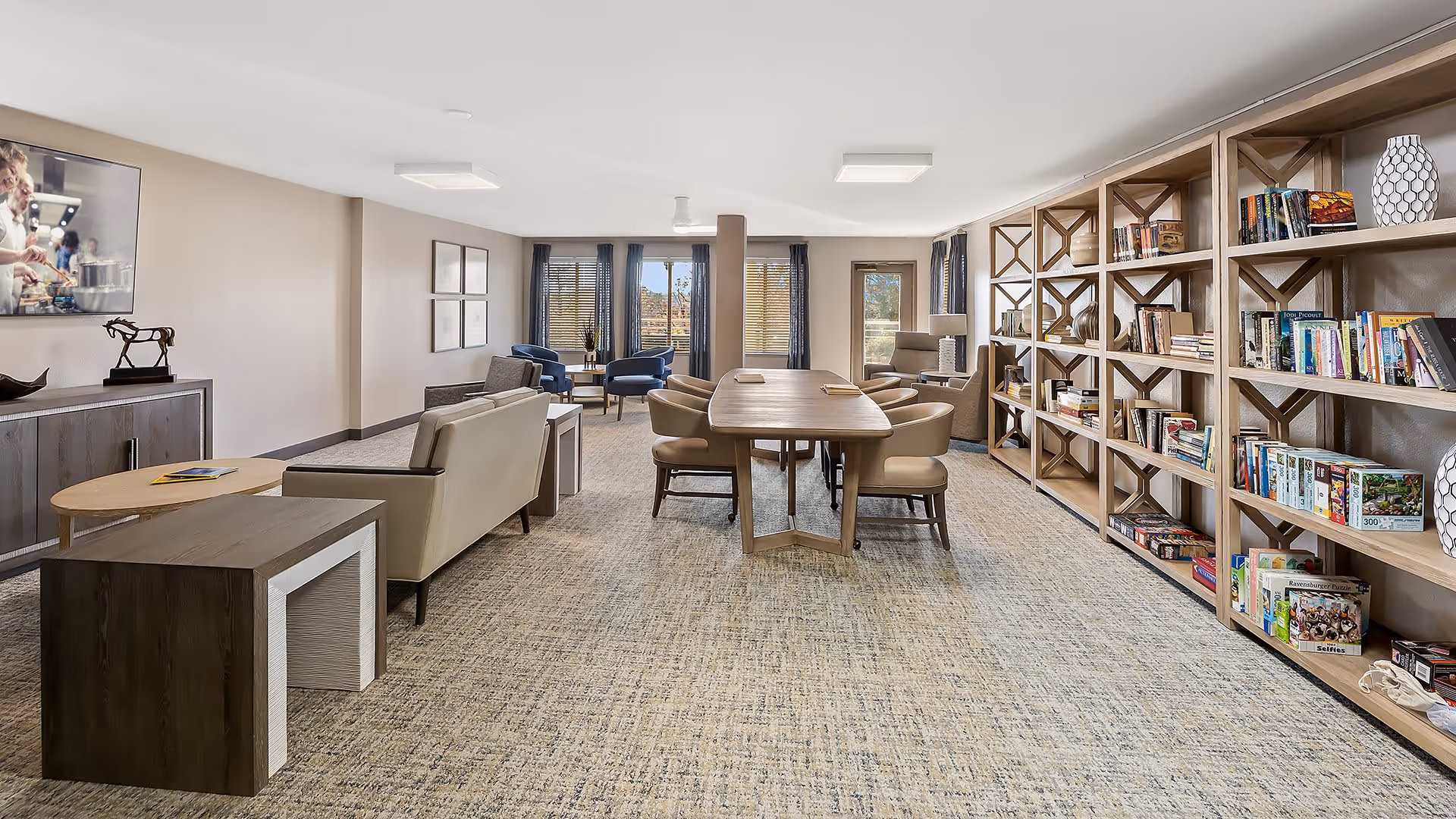 A spacious and well-lit common area in a senior living facility featuring a large wooden table with chairs in the center, comfortable seating including sofas and armchairs, a large bookshelf filled with books and games along one wall, and large windows with curtains allowing natural light to fill the room.