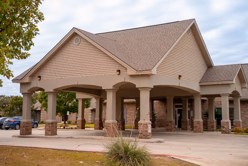 Entrance area of a senior living facility with a covered drop-off zone supported by columns, surrounded by a parking lot and some greenery under a cloudy sky.