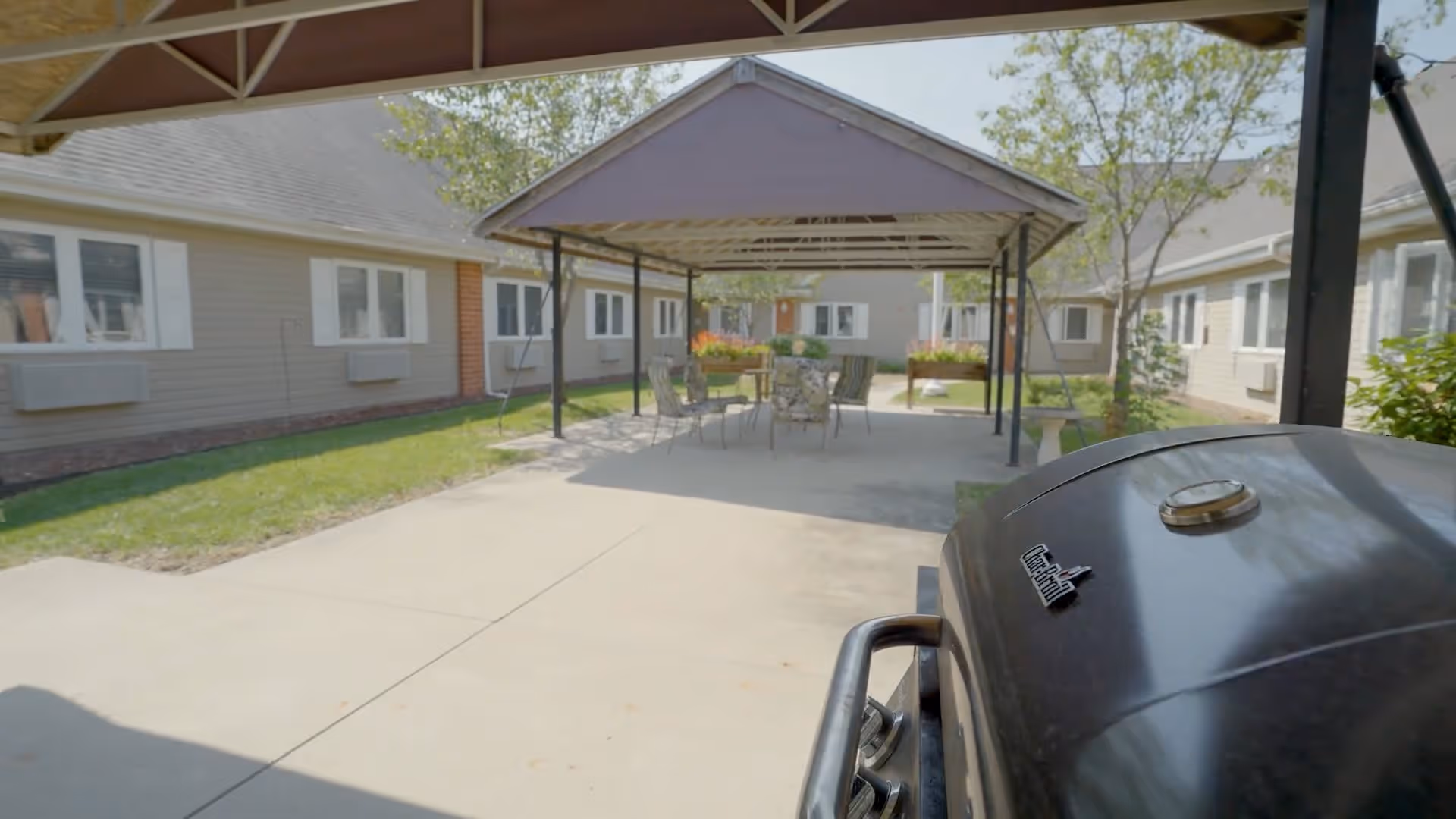 Outdoor patio area at Regency Assisted Living with a covered seating area featuring a table and chairs, surrounded by single-story buildings with windows and greenery.