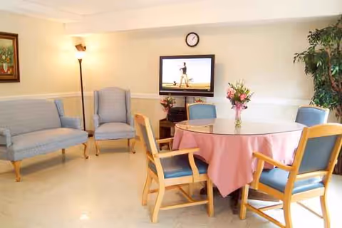 Senior living facility lounge with round table, pink tablecloth, blue chairs, TV, and plants.