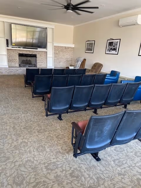 A senior living facility common room with rows of black theater-style chairs facing a wall-mounted flat screen TV above a fireplace. The room has beige walls, a ceiling fan, framed black and white photos on the wall, and a few armchairs in the corner.