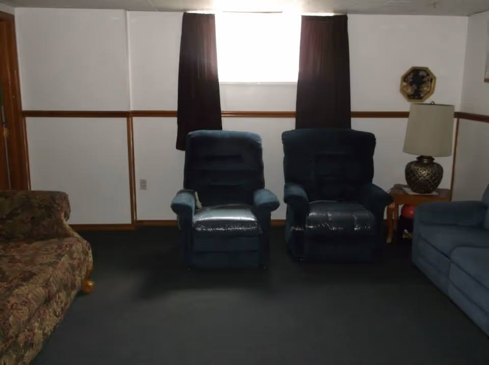 Two dark recliner chairs and sofas in a modest living room with a small curtained window and a side table lamp.
