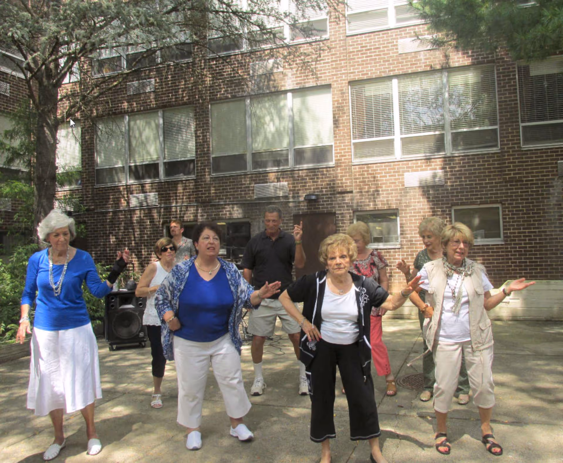 A group of elderly people standing outdoors in a courtyard area in front of a brick building, engaging in a group activity or exercise. There are trees providing shade and a speaker visible in the background.