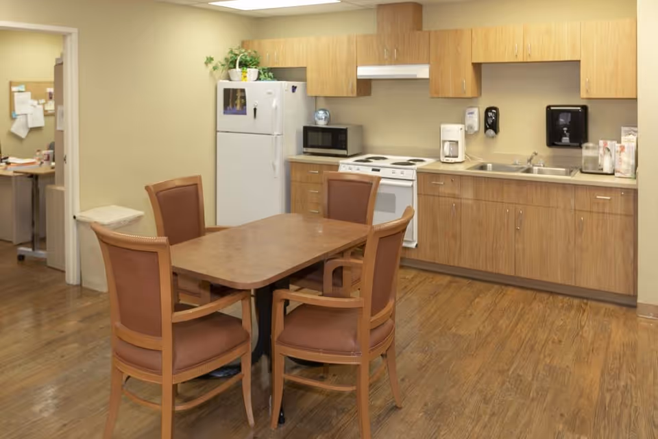 A small kitchen area with wooden cabinets, a white refrigerator, microwave, stove, coffee maker, and sink. In front of the kitchen is a rectangular wooden table with four cushioned chairs. The floor is wood, and there is an open doorway leading to an office space with a desk and bulletin board.