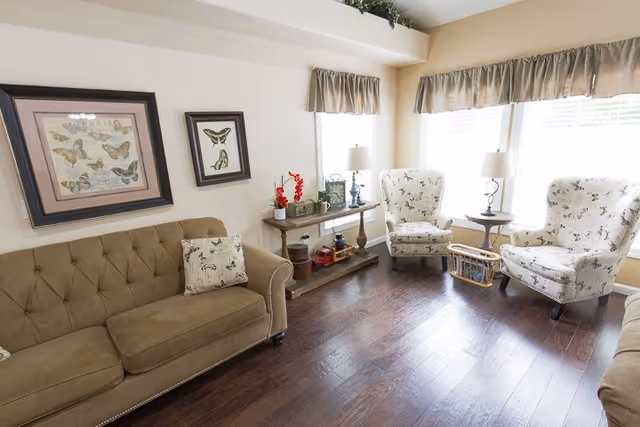 A cozy living room with a brown tufted sofa, two patterned armchairs, a wooden console table with decorative items, and two windows with beige valances letting in natural light. The floor is dark hardwood.