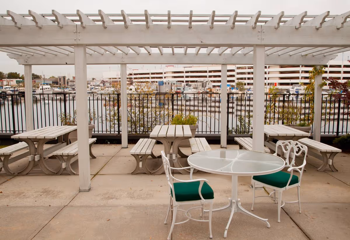Outdoor patio area with white pergola overhead, featuring white picnic tables with benches and a round glass-top table with two white chairs that have green cushions. In the background, there is a black metal fence, water with boats docked, and a multi-level parking structure.