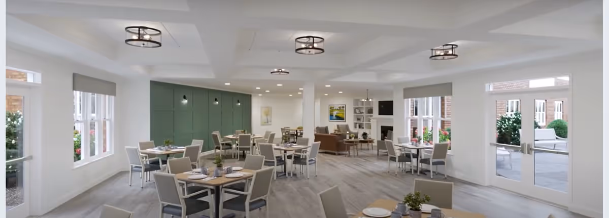 A spacious and bright senior living dining area with multiple tables and chairs arranged neatly. The room features large windows and glass doors allowing natural light to fill the space, light wood flooring, modern ceiling lights, and a cozy seating area with sofas and a fireplace in the background. Outside the glass doors, a patio with outdoor seating and greenery is visible.