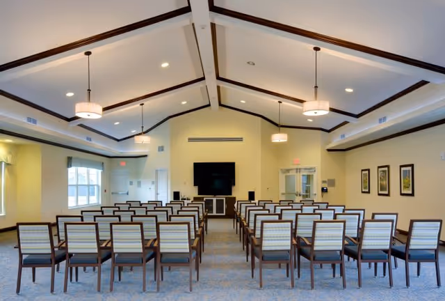 A bright multipurpose meeting room with rows of chairs facing a wall-mounted TV under a vaulted ceiling.