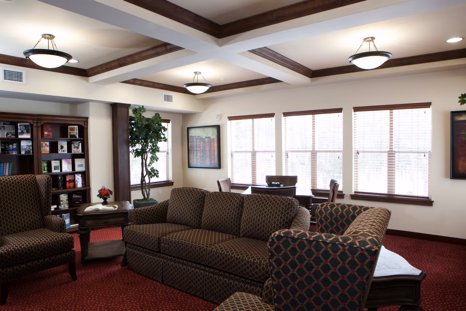 A cozy living room area in a retirement facility with patterned upholstered sofas and armchairs, a wooden coffee table with a small flower arrangement, a bookshelf filled with books, large windows with blinds letting in natural light, and framed artwork on the walls. The ceiling features wooden beams and modern light fixtures.