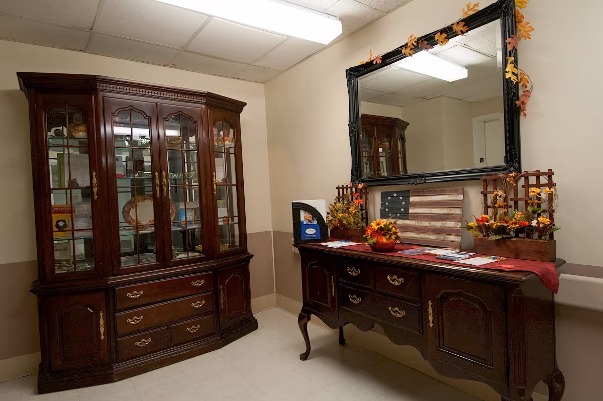 Interior room with dark wooden furniture including a glass-front cabinet and a sideboard with drawers. The sideboard is decorated with autumn-themed floral arrangements, a small American flag artwork, and a large black-framed mirror above it. The walls are beige with a two-tone paint design, and the ceiling has fluorescent lighting.
