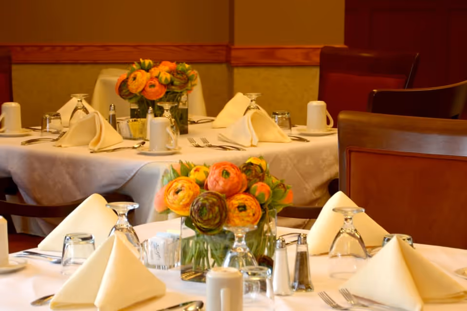 A dining room with tables set for a meal, featuring white tablecloths, folded beige napkins, glassware, coffee cups, silverware, salt and pepper shakers, and floral centerpieces with orange and yellow flowers.