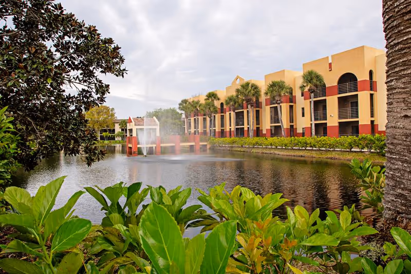 Tan-and-red multi-story assisted living building beside a pond with a fountain, palm trees, and foreground greenery.