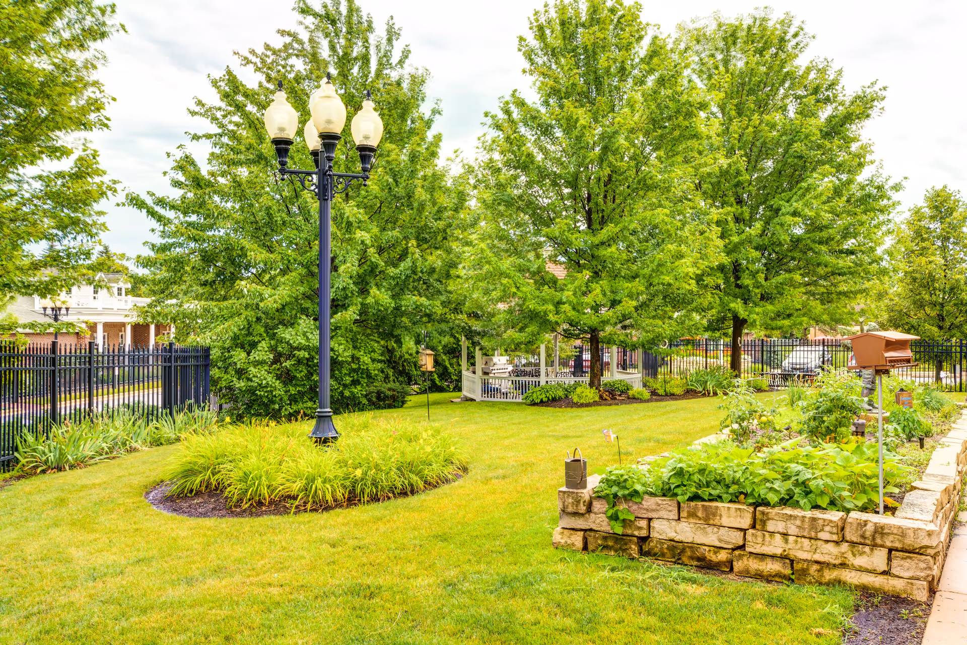 A well-maintained outdoor garden area with lush green grass, several trees, a black metal fence, a vintage-style street lamp, and a raised stone planter box filled with various plants and vegetables. There is also a small wooden birdhouse on a pole and a paved walkway on the right side.