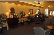 A warmly lit interior lounge area with upholstered chairs and small tables, decorative plants, a large mirror above a wooden sideboard, and wall-mounted lamps. The space has carpeted flooring and chandeliers hanging from the ceiling.