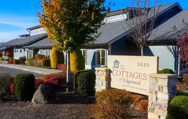 Exterior view of The Cottages at Edgewood senior living facility with a stone sign displaying the community name and address 2510, surrounded by landscaped bushes and trees under a clear blue sky.