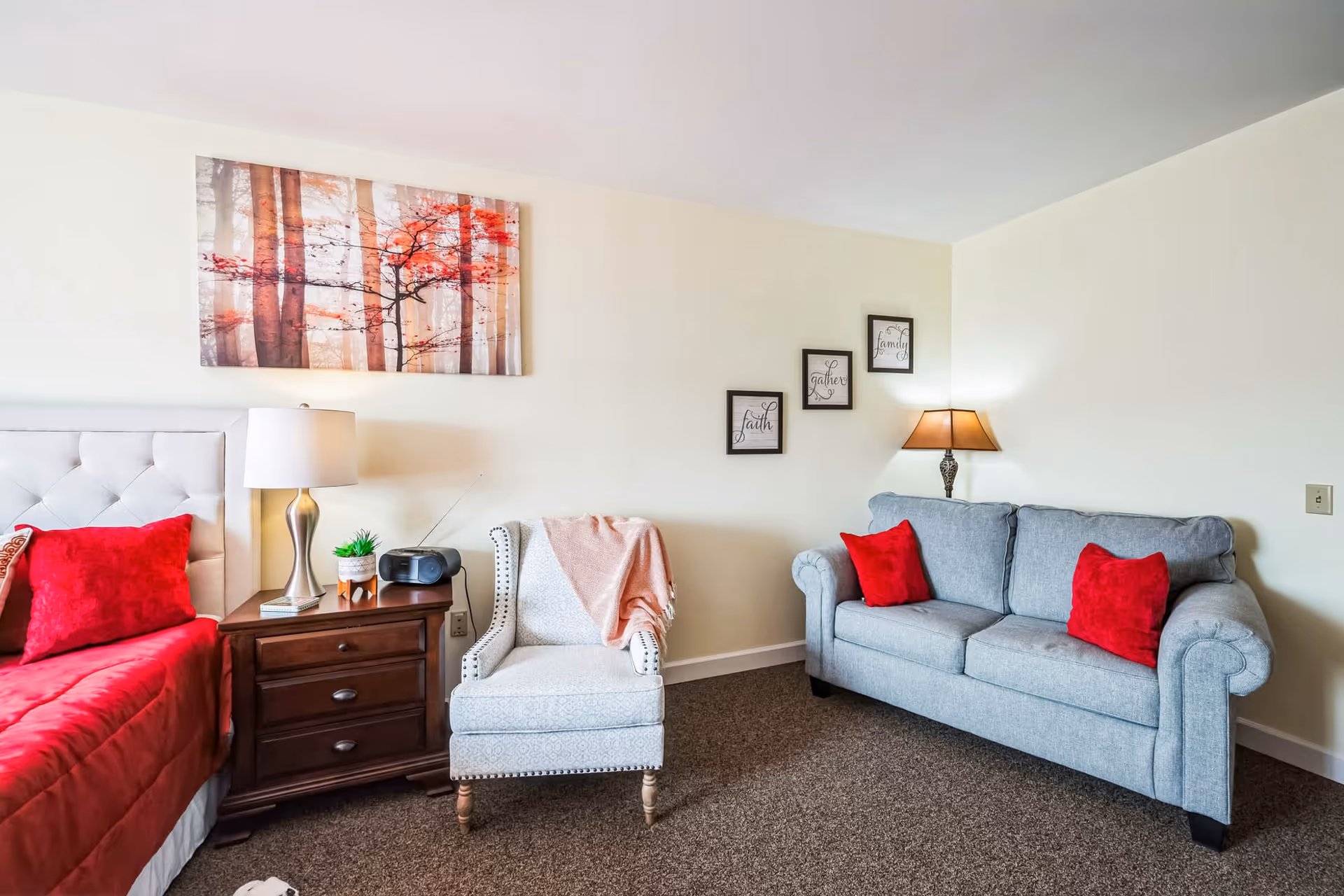 A cozy living space featuring a gray upholstered armchair with a pink throw draped over it, a wooden nightstand with a lamp, a small plant, and a radio, a gray couch with red pillows, and a bed with red bedding and pillows. The walls are decorated with a large painting of trees with red leaves and three small framed quotes.