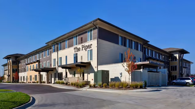 Exterior view of The Piper Assisted Living and Memory Care facility, a modern three-story building with beige and brown walls, multiple windows, and a clear blue sky above.