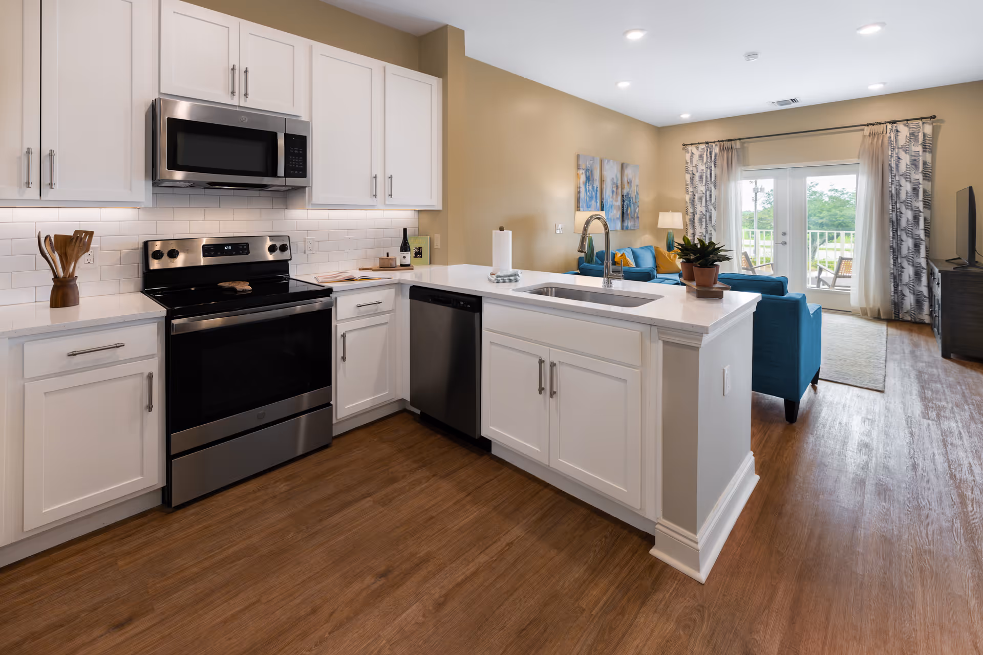 Modern kitchen with white cabinets, stainless steel appliances including a stove, microwave, and dishwasher, a white countertop island with a sink and a potted plant. In the background, a living area with blue furniture, a TV, and glass doors leading to an outdoor balcony.