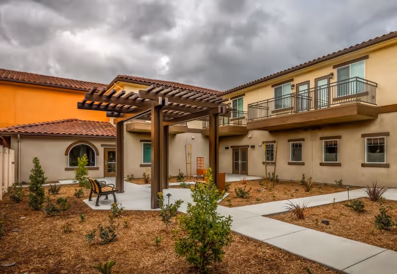 Outdoor courtyard area of a senior living facility with a wooden pergola, benches, young plants, and a two-story building with balconies and windows under a cloudy sky.