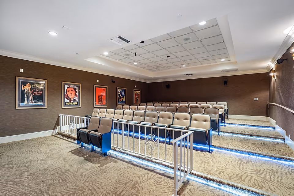 Interior view of a small theater room with tiered seating featuring beige cushioned chairs. The room has brown walls adorned with framed vintage movie posters, a carpeted floor with a leaf pattern, and blue LED lighting along the steps. The ceiling has recessed lighting and acoustic panels.