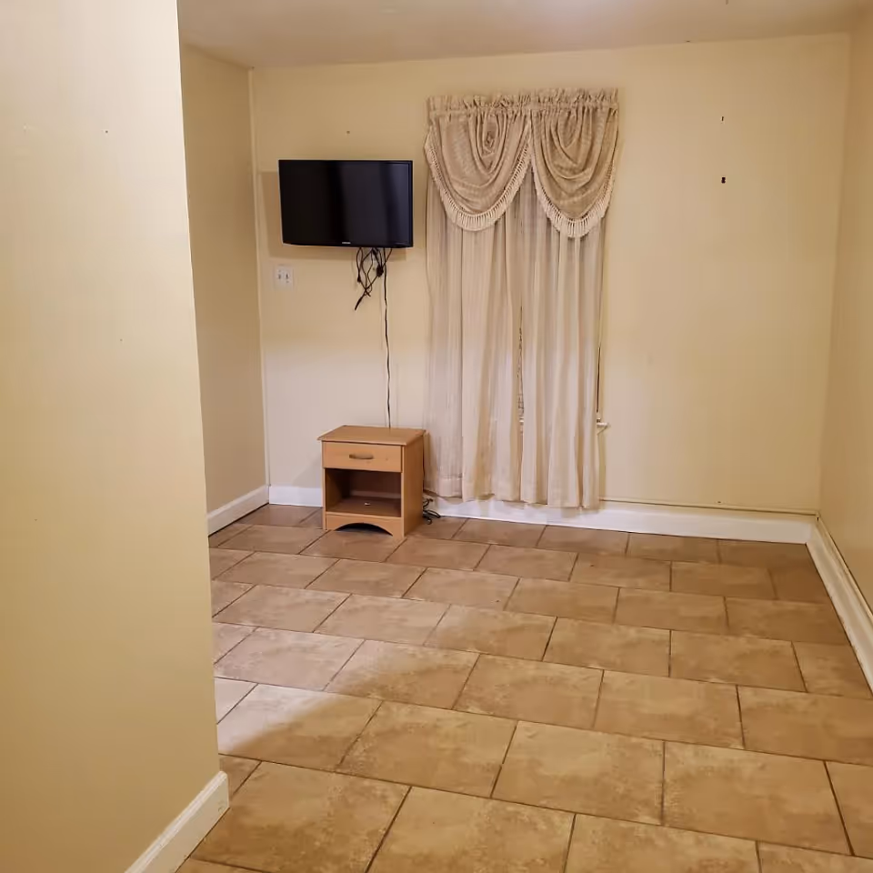 Small mostly empty interior room with tiled floor, a wall-mounted TV above a small wooden nightstand, and a curtained window.